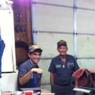 Two people in work shirts and caps smile while standing in a garage workspace.