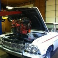 A red engine is being lowered by a hoist into the engine bay of a white classic car inside a garage.