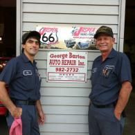 Two people in matching navy work uniforms stand in front of a George Barton Auto Repair sign on a grey siding wall.