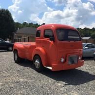 A bright orange vintage cab-over truck parked on a gravel lot under a partly cloudy sky.