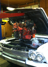 An orange car engine being lifted from the engine bay of a white car by a red engine hoist inside a garage.