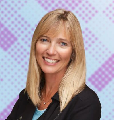 Blonde woman with blue eyes smiles, wearing black jacket, against patterned background.