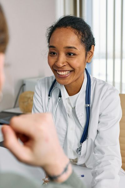 Doctor smiling, talking with a patient. She's wearing a white coat and has a stethoscope. Office setting.