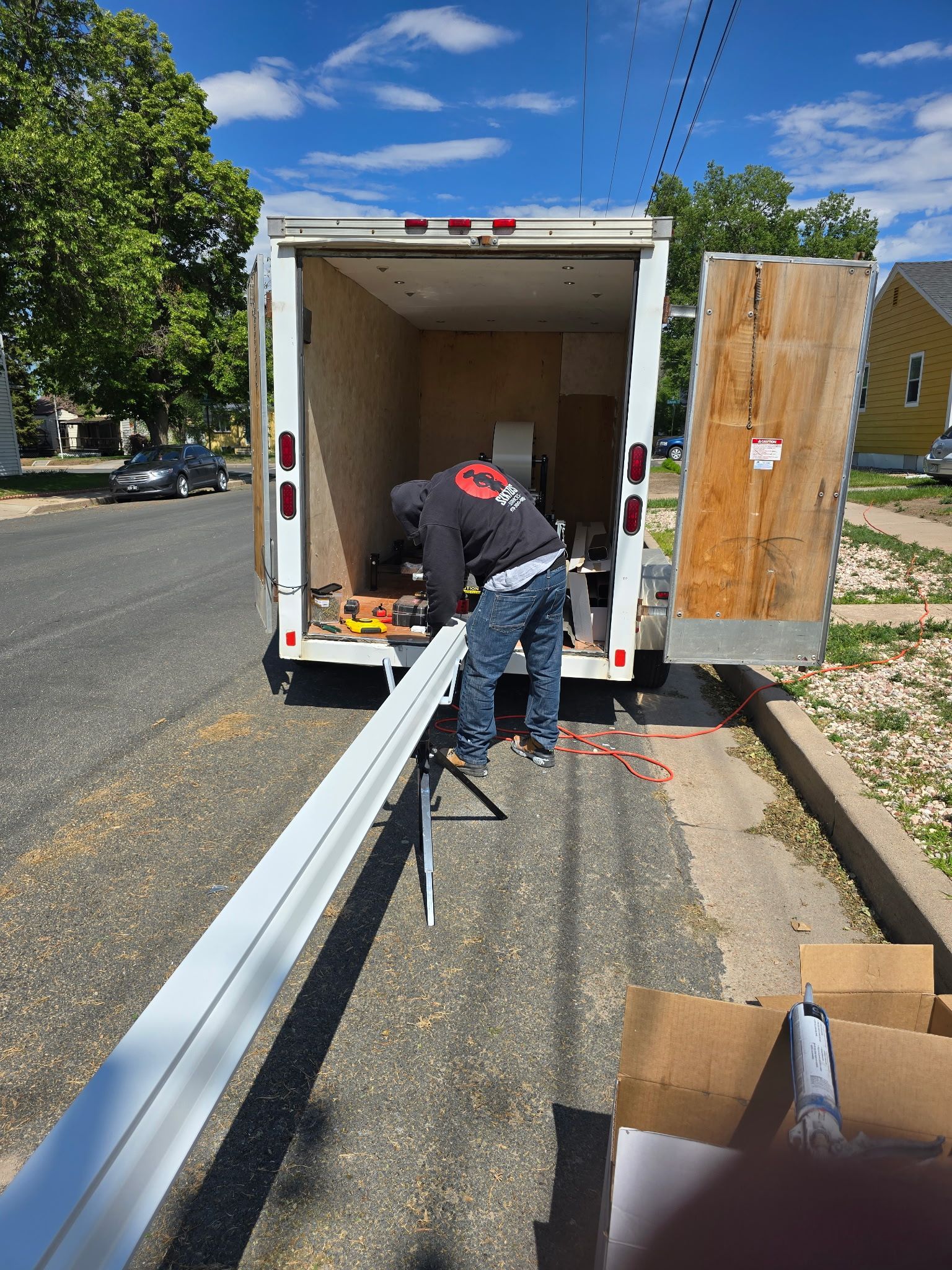 Person working on a long, white metal piece near an open trailer, on a sunny day.
