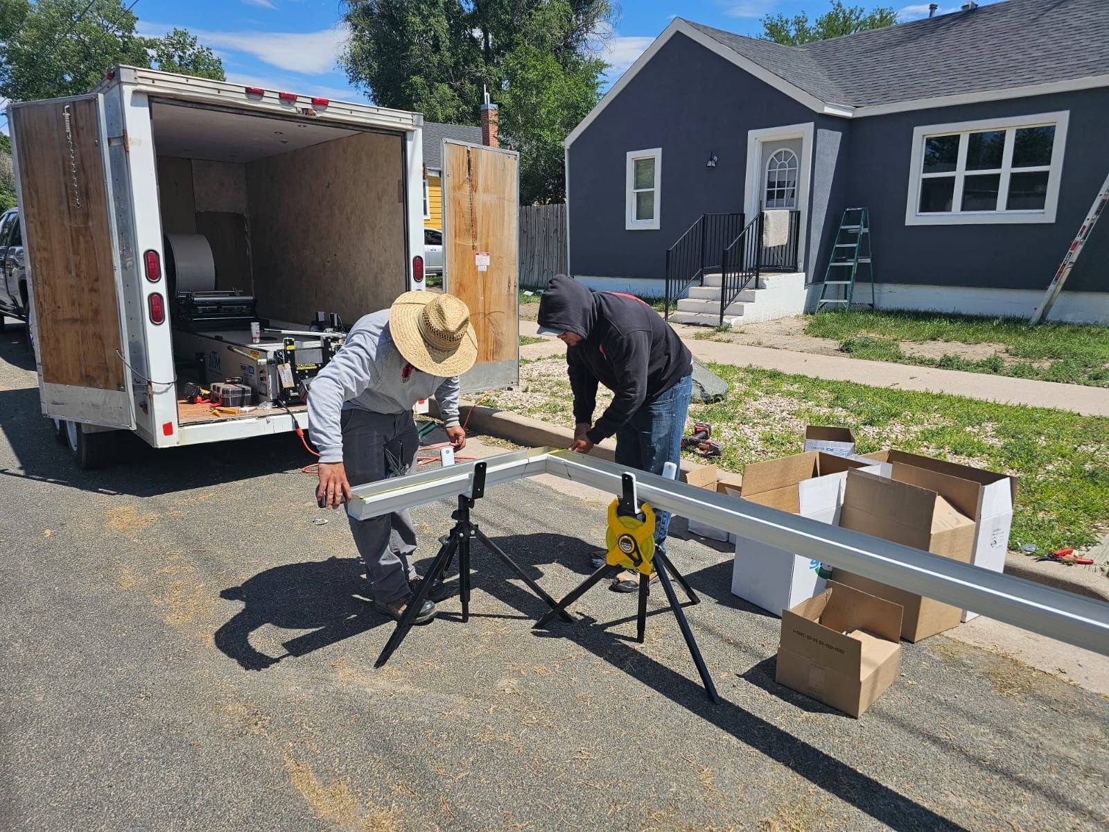 Two people cutting metal siding on a street next to a trailer and a blue house.