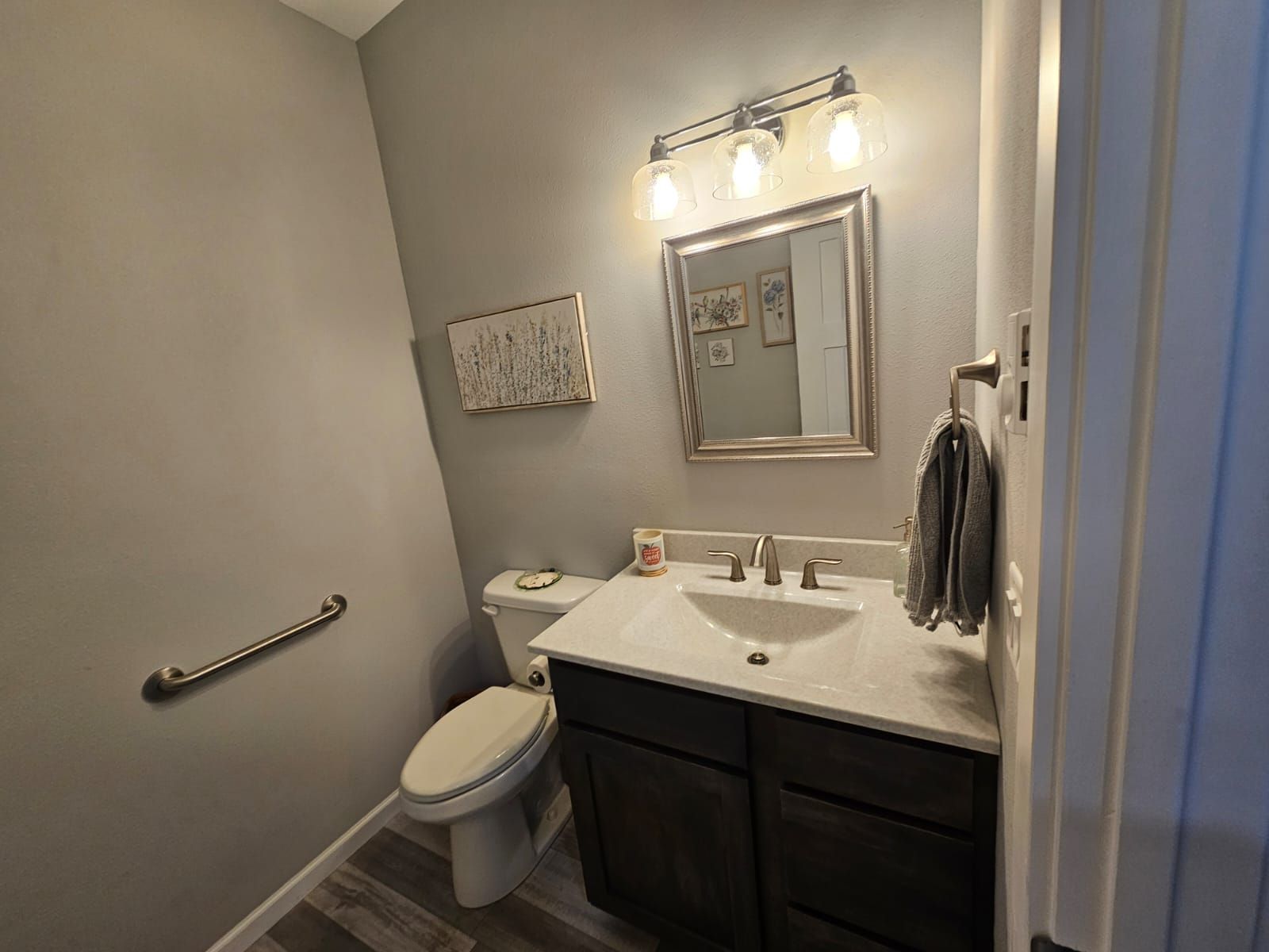 Small bathroom with a toilet, sink, mirror, and a handrail. Gray walls and dark brown cabinet.
