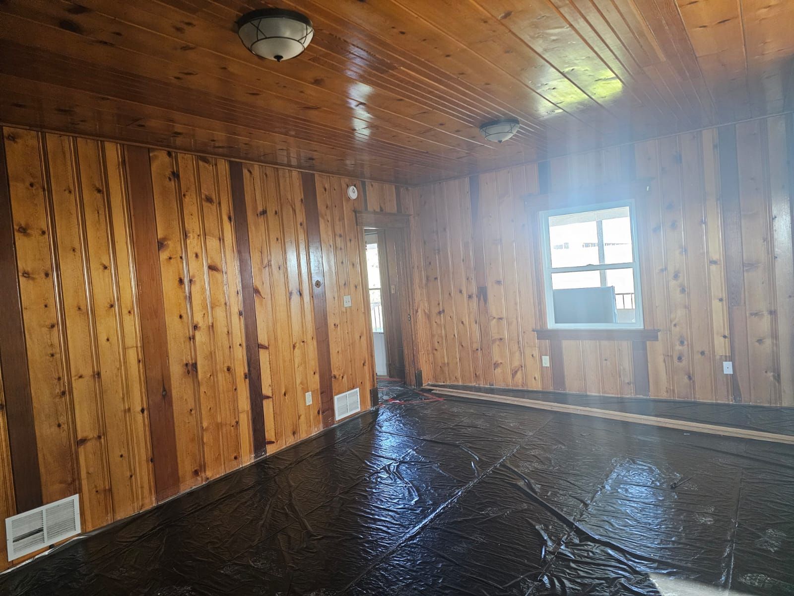 Room with wood paneling on walls and ceiling, a window, and a dark covering on the floor.