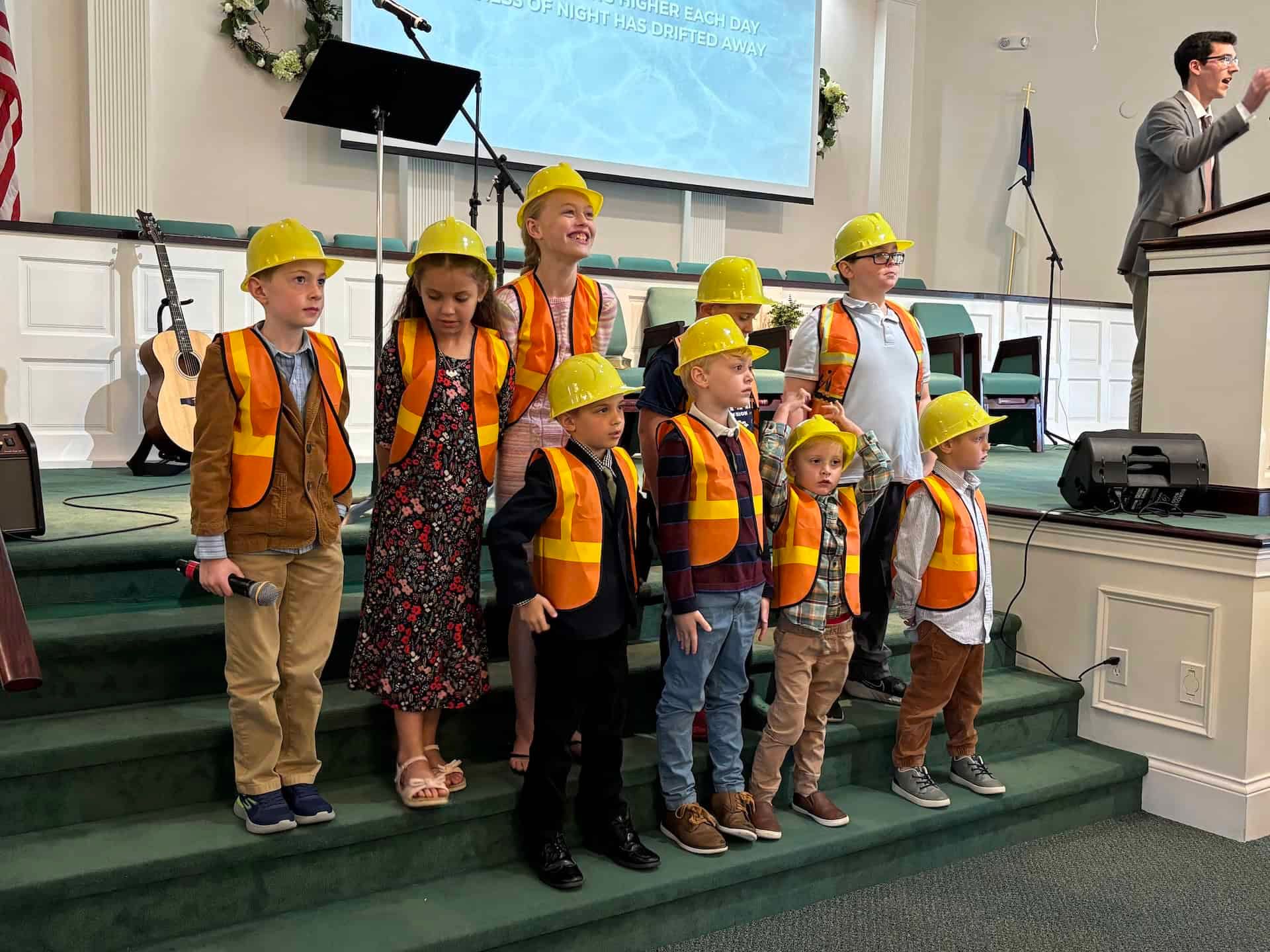 A group of children wearing hard hats and safety vests are standing on a stage.