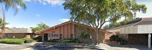 Low-angle view of a tan-colored building with a rock and wood façade, lush green foliage, and a large tree.