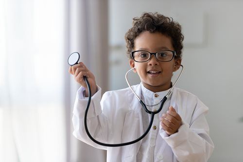 Boy in a lab coat and glasses holds a stethoscope, smiling.