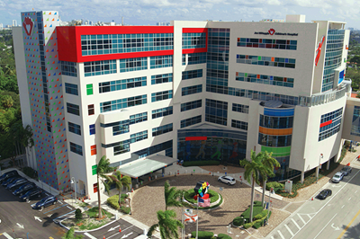 Miami Children's Hospital exterior with red, white, and blue building, climbing wall, and palm trees.