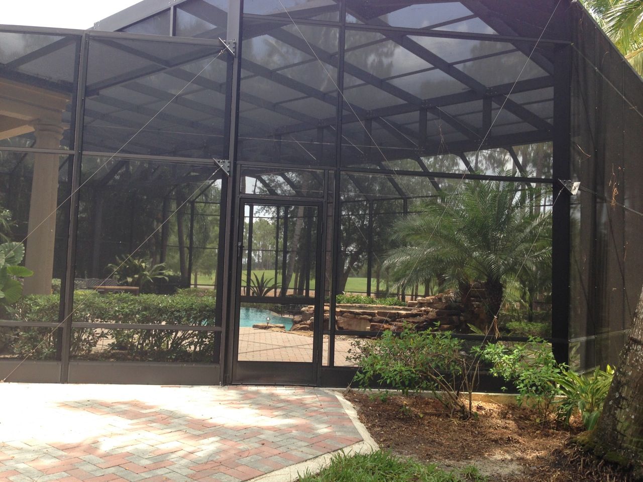 Black screened-in pool enclosure with a door, plants, and trees in the background.