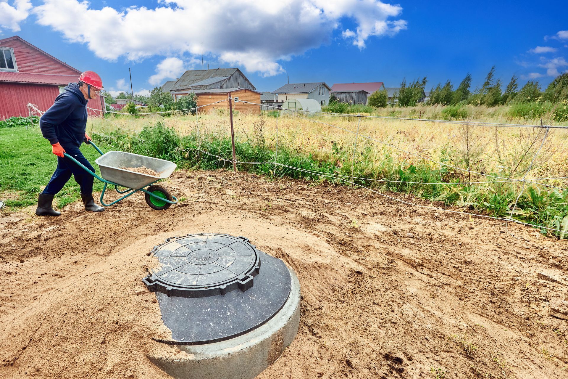Worker in hard hat pushing wheelbarrow near a newly installed septic tank cover in a sandy backyard