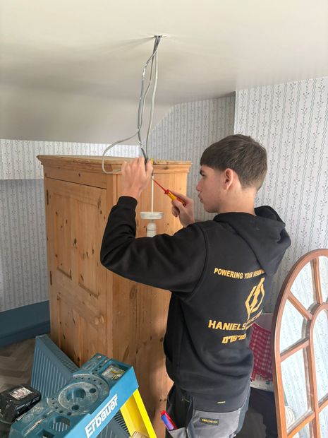 Person wiring a wooden cabinet to the ceiling in a room with tools and a blue ladder below.