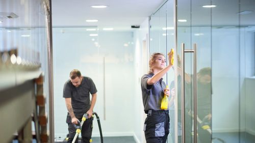 A man and a woman are cleaning a glass door in an office.