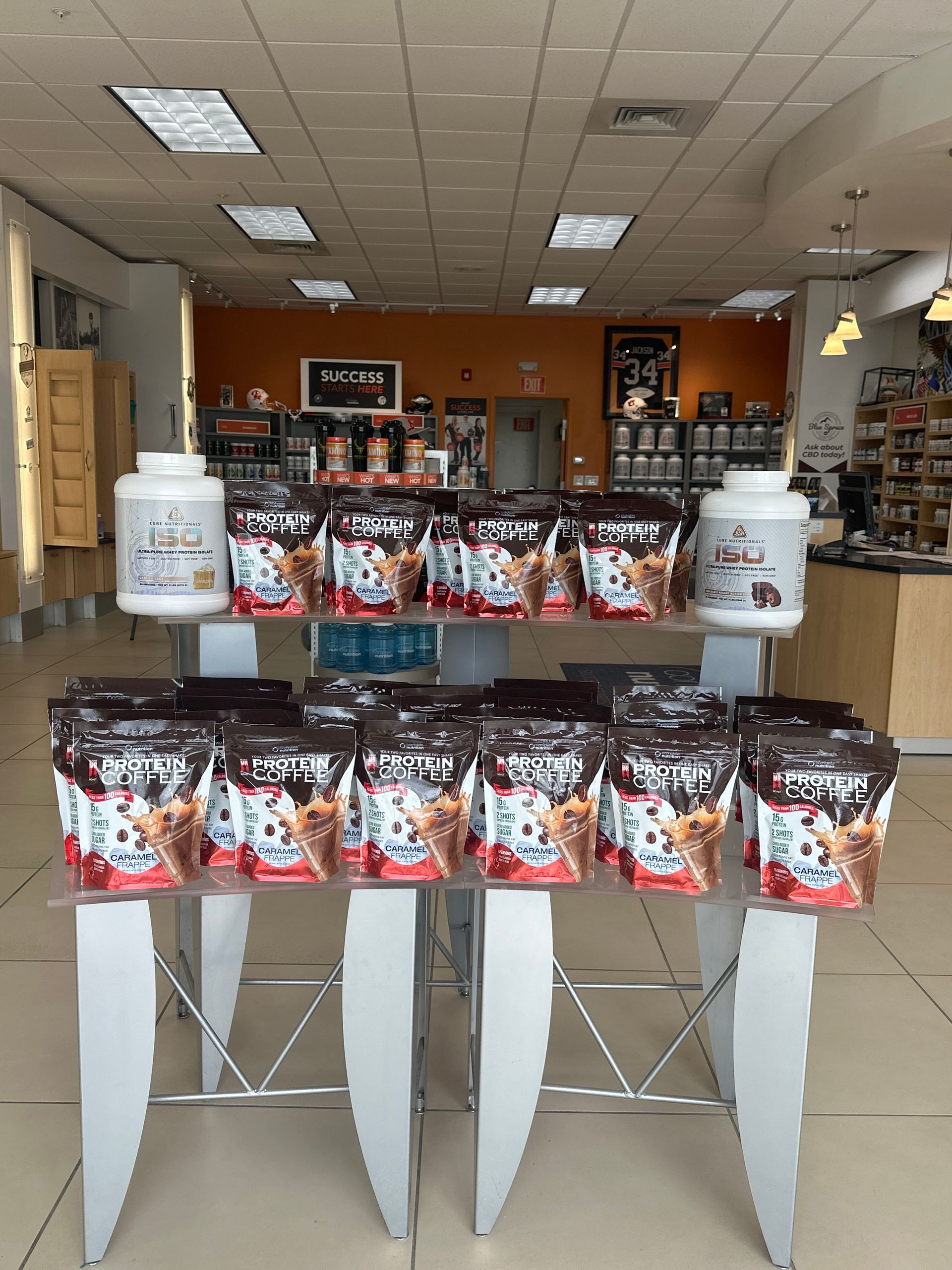 A store display featuring protein powder containers and packaged granola bars, with overhead lighting.