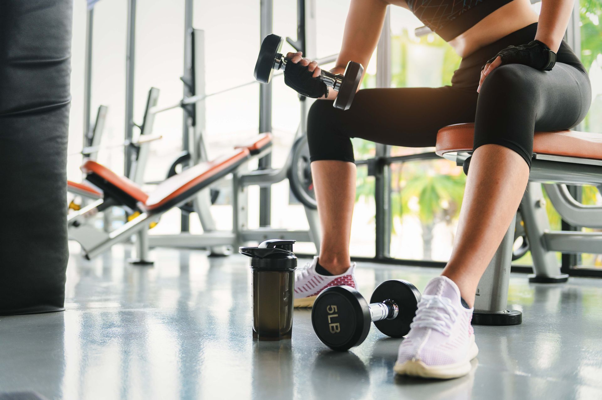 Person seated at a gym bench, holding dumbbells, ready to exercise, with a protein shaker bottle.