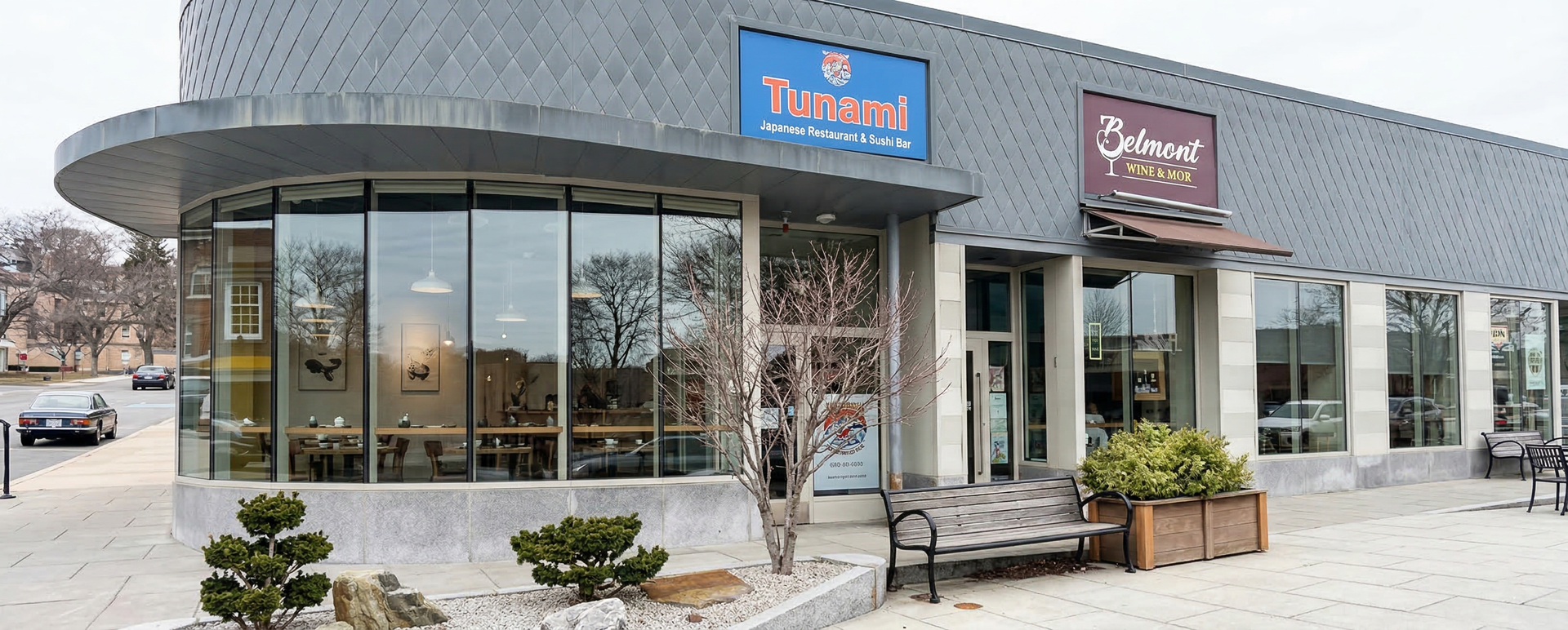 A modern, curved storefront with glass windows, a dark facade, and a bench outside on a sidewalk.