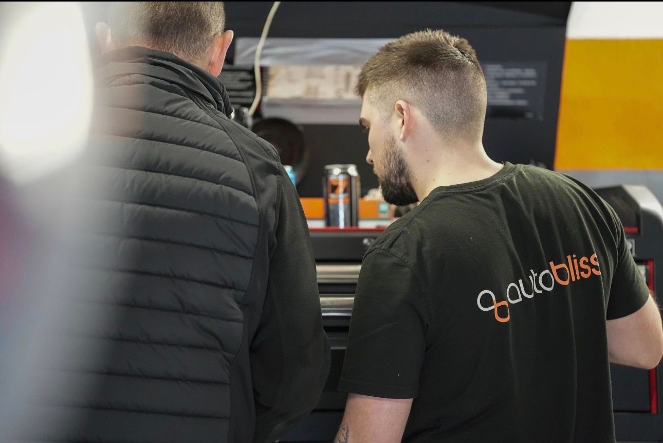 Two people stand in a garage workshop, focusing on equipment on a workbench with a soda can nearby.