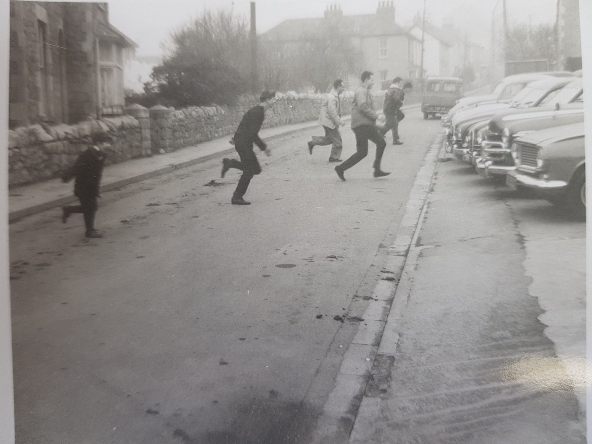 A black-and-white photo of several people running across a street lined with parked cars and stone walls.