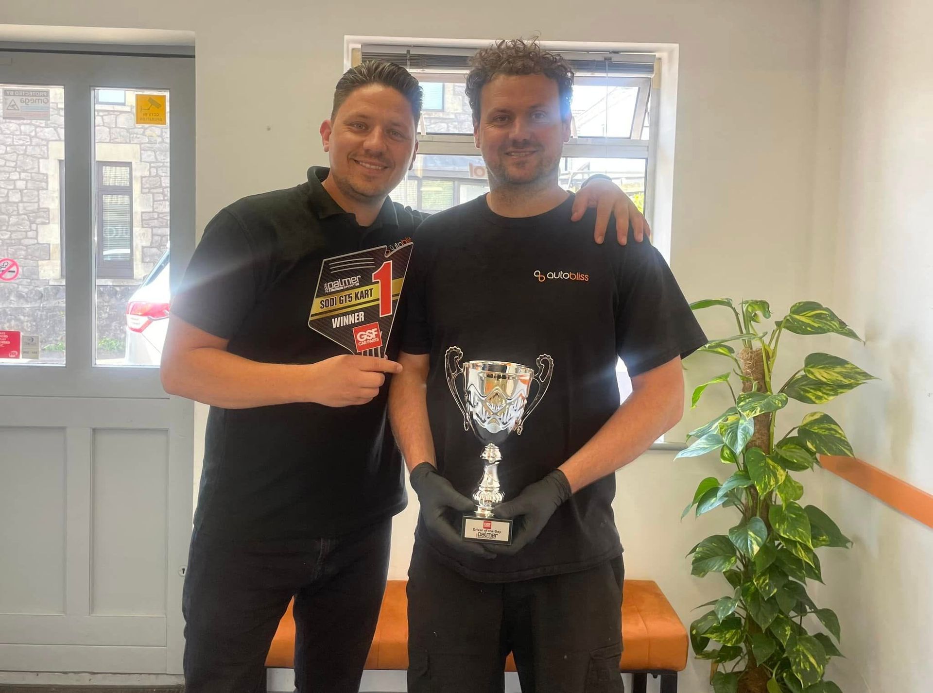 Two smiling individuals stand side-by-side in a room, with one holding a silver trophy while wearing black gloves.