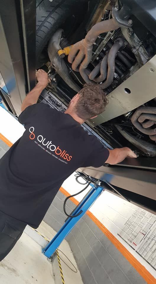 A mechanic wearing an Auto Bliss black t-shirt works under a car raised on a hydraulic lift in a garage.