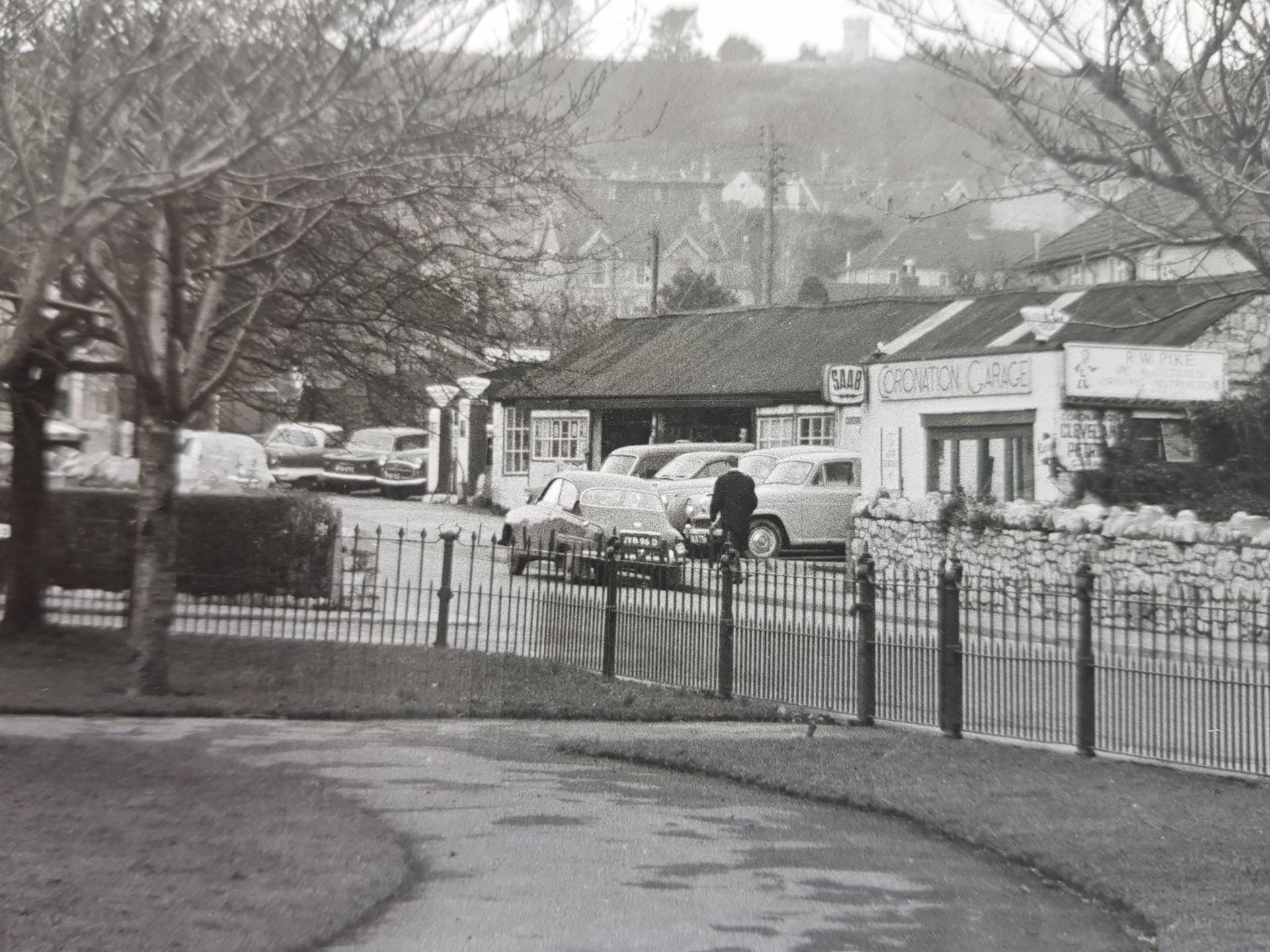 A black-and-white view of a roadside garage with parked cars, separated from a foreground park path by a metal fence.