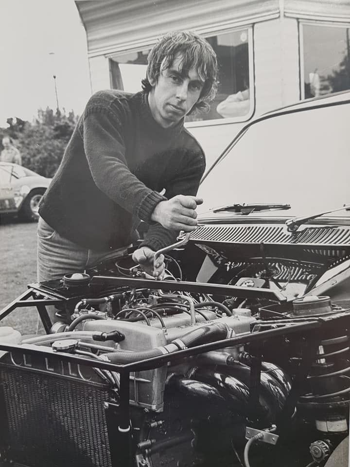 A person in a dark sweater works on the open engine of a race car in a black-and-white outdoor setting.