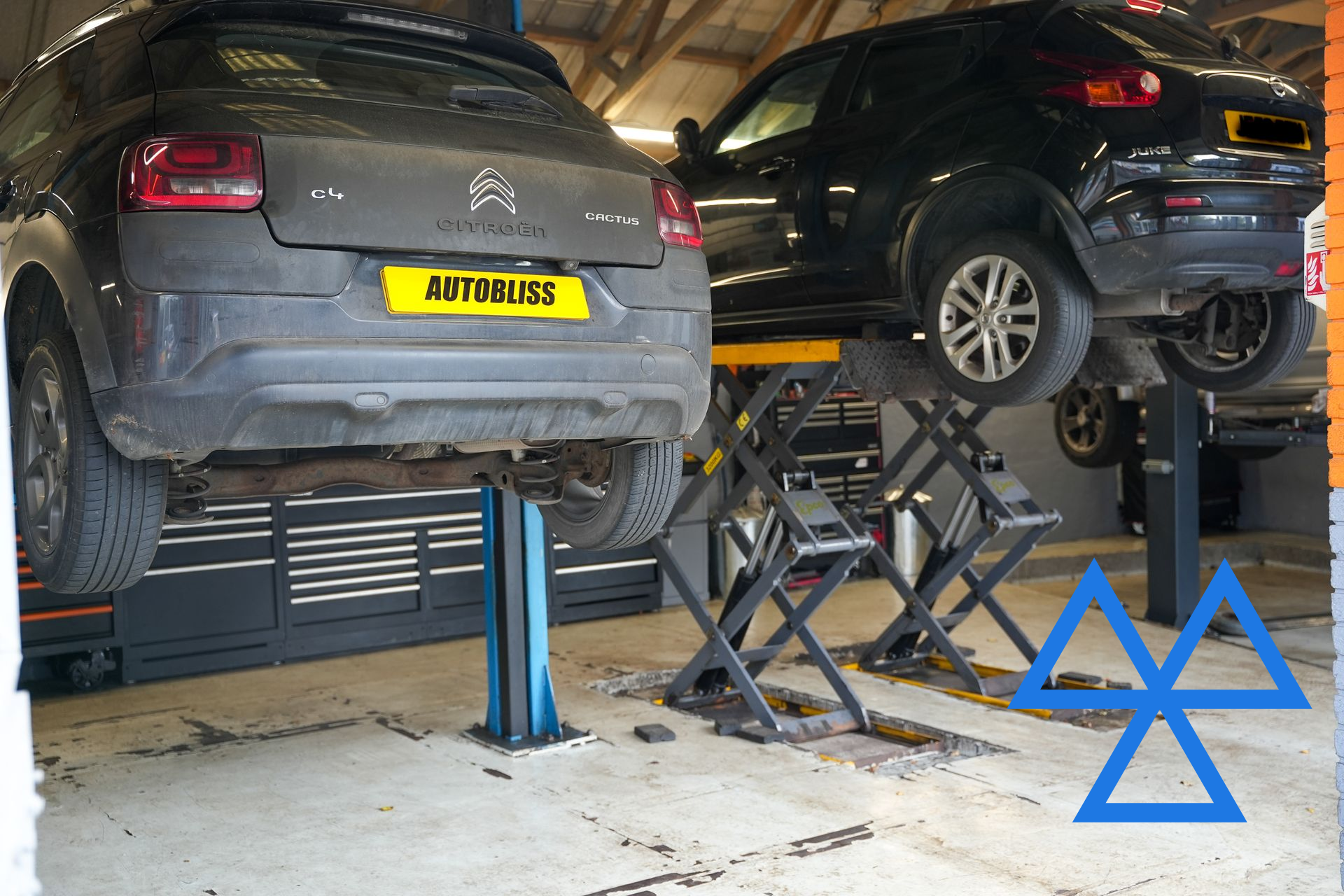 Two cars on hydraulic lifts inside a garage, with a blue MOT test center symbol superimposed in the corner.