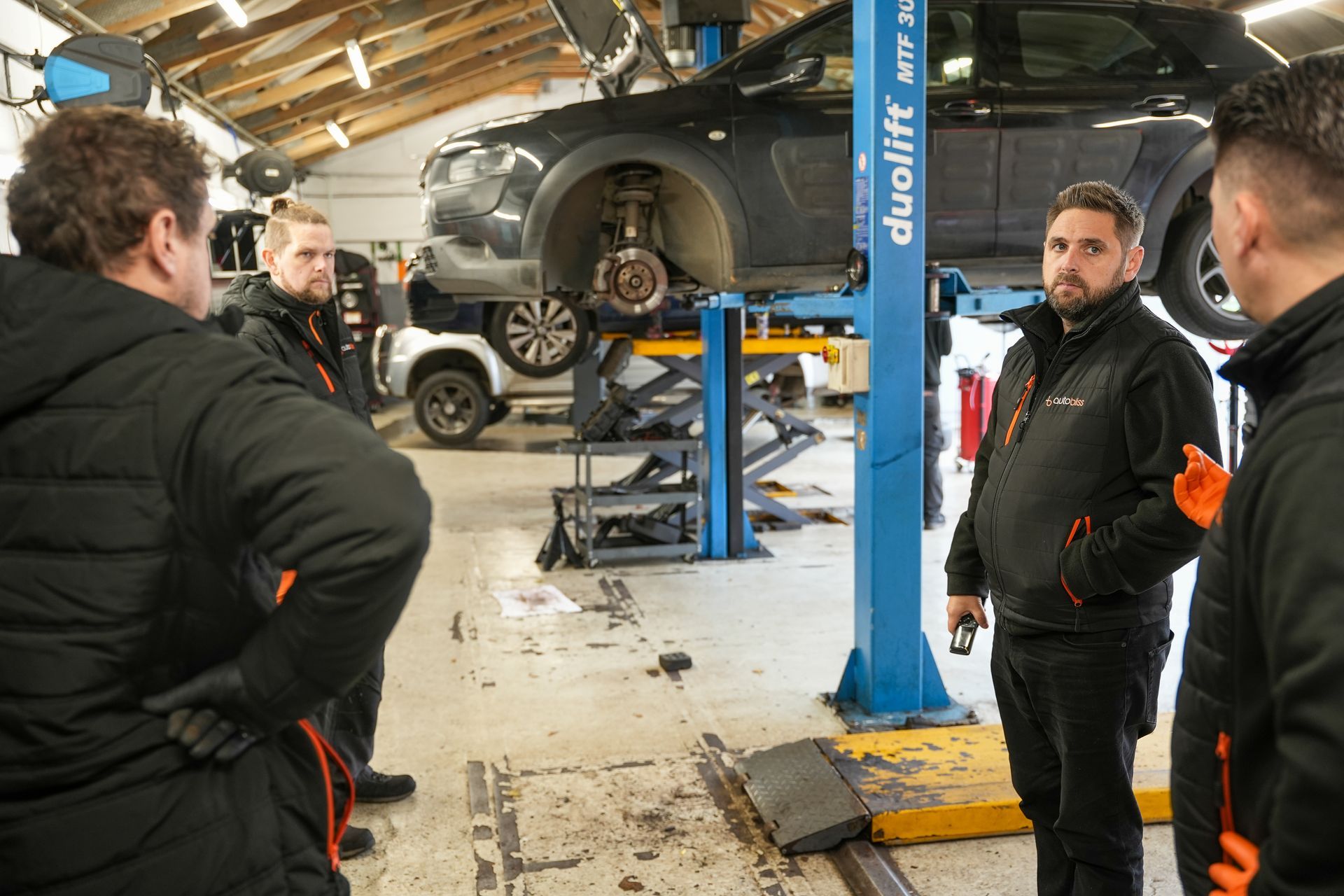 Four mechanics in uniform stand in a workshop discussing a car raised on a blue lift.