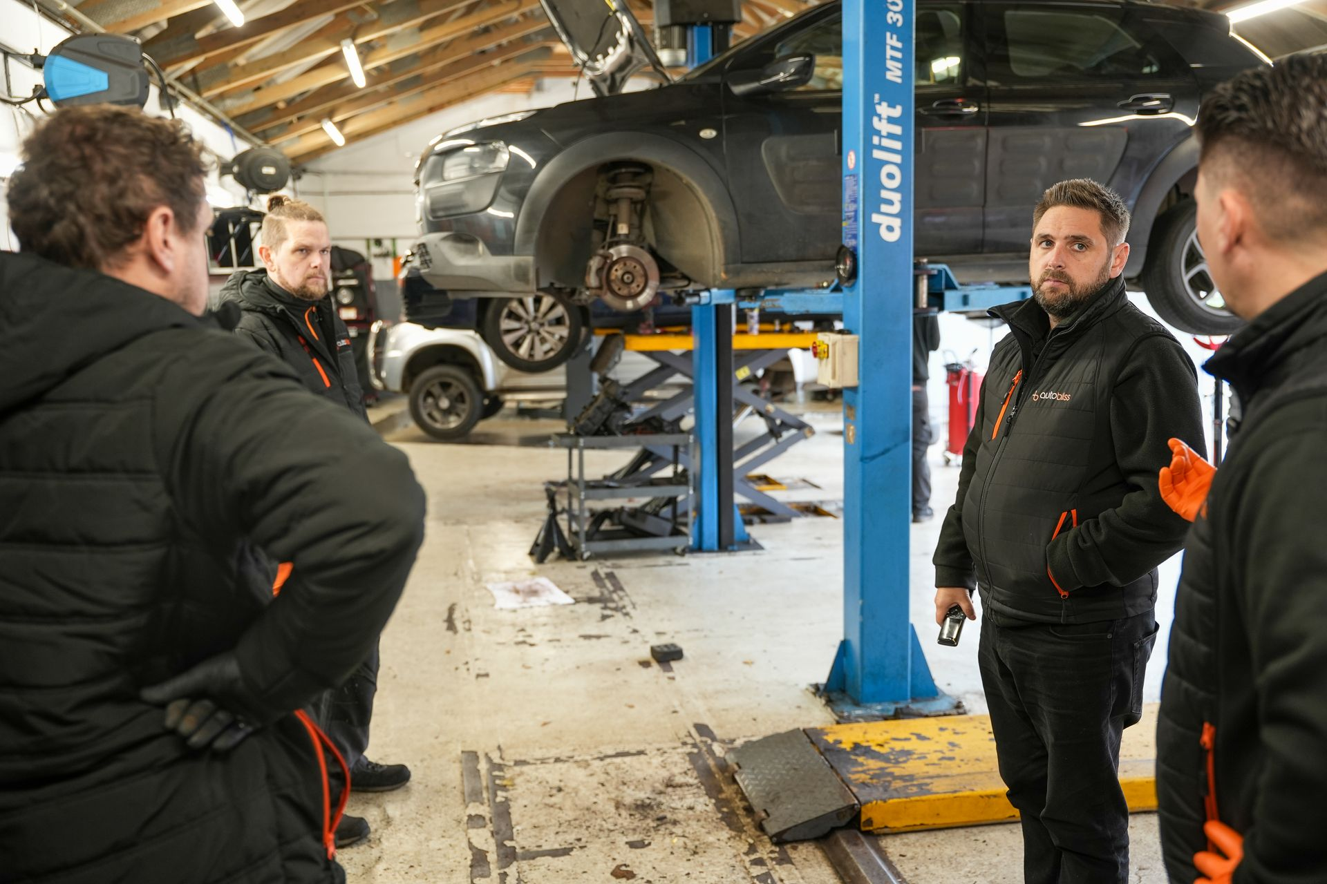 Four mechanics in work uniforms stand in a repair shop discussing a vehicle raised on a blue lift.