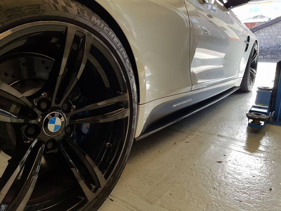Close-up side view of a white BMW sedan featuring dark alloy wheels, carbon fiber side skirts, and a shop floor setting.