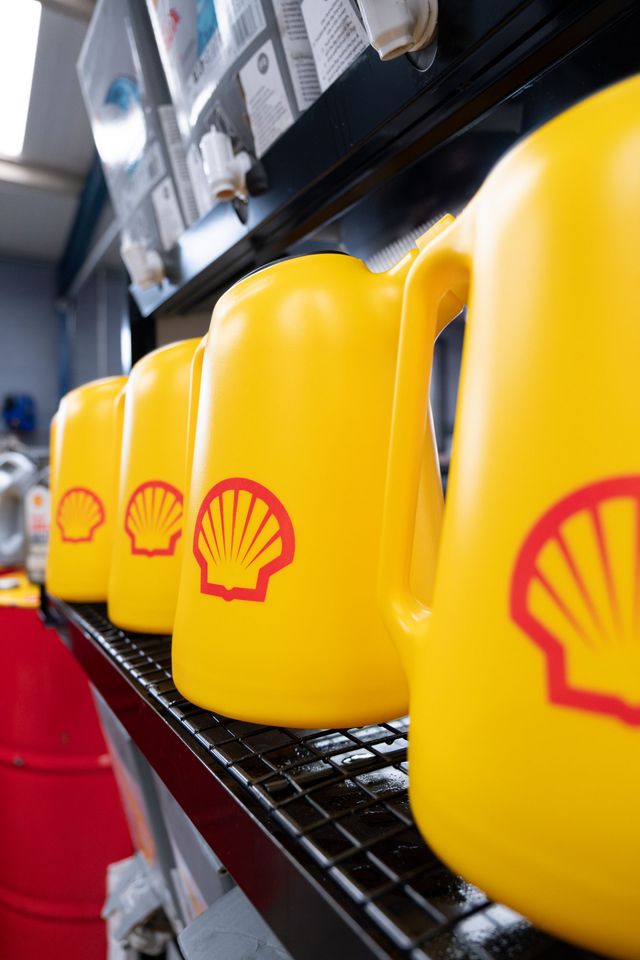 A row of bright yellow Shell oil jugs with red logos sits on a black shelf in an industrial or automotive setting.