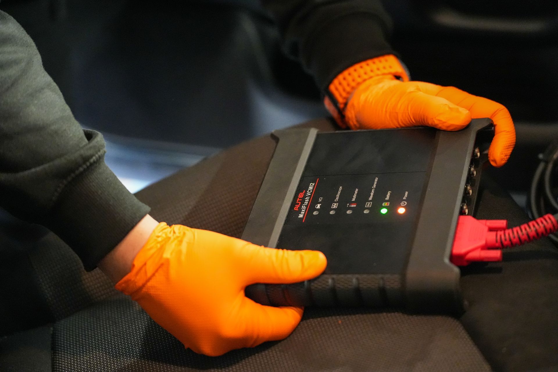 A person wearing orange gloves holding a car diagnostic scanner device with glowing status lights.