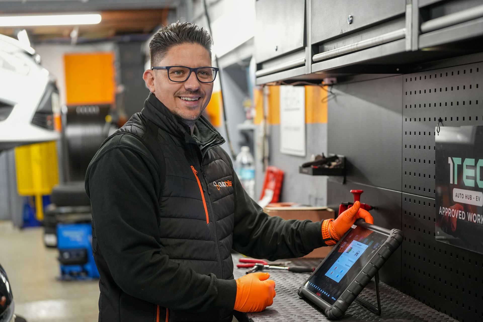 A technician in a garage wears orange gloves while using a handheld diagnostic tablet at a workbench.
