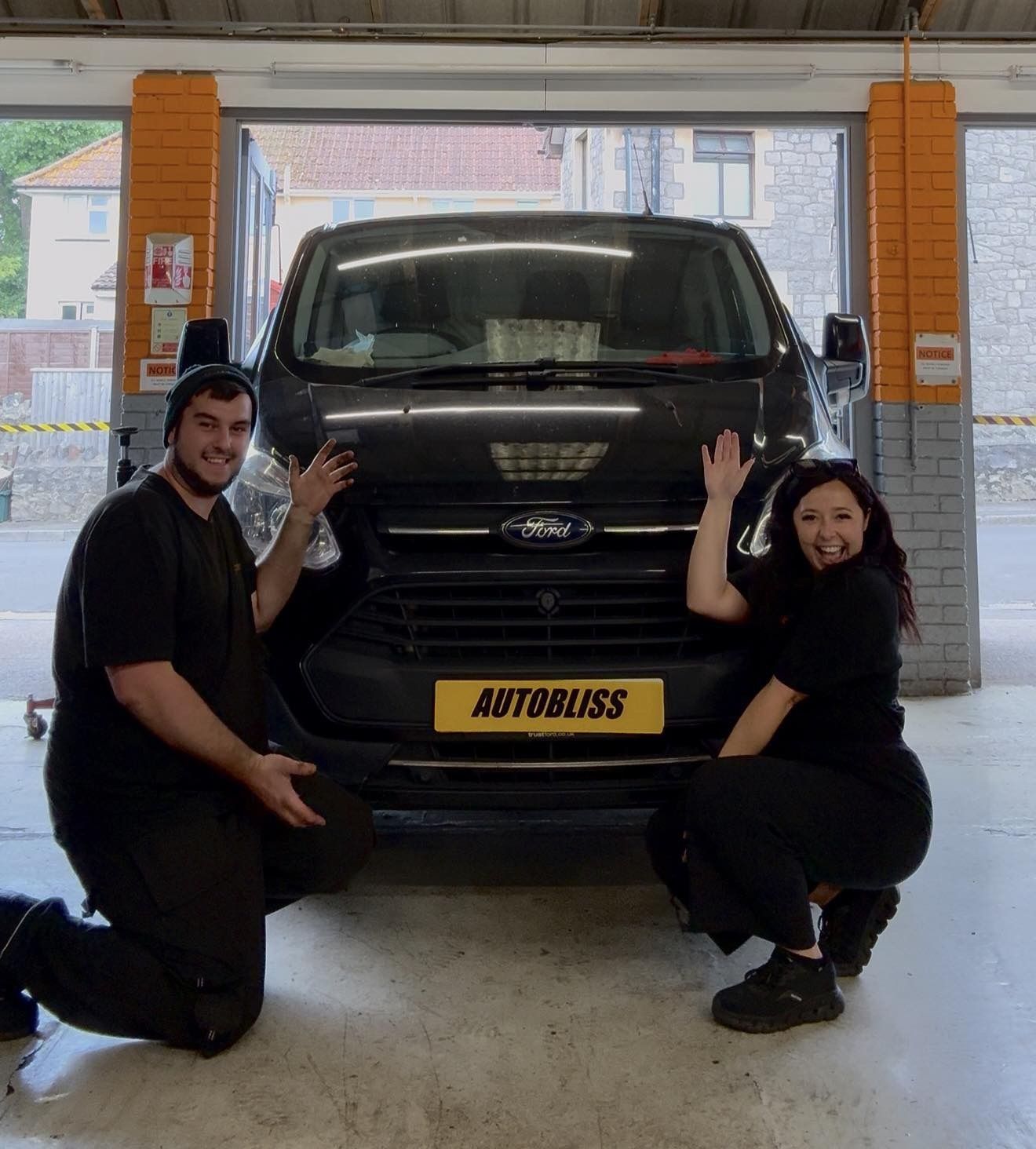 Two people in dark clothing pose on their knees in a garage, smiling and waving beside a black Ford van with 