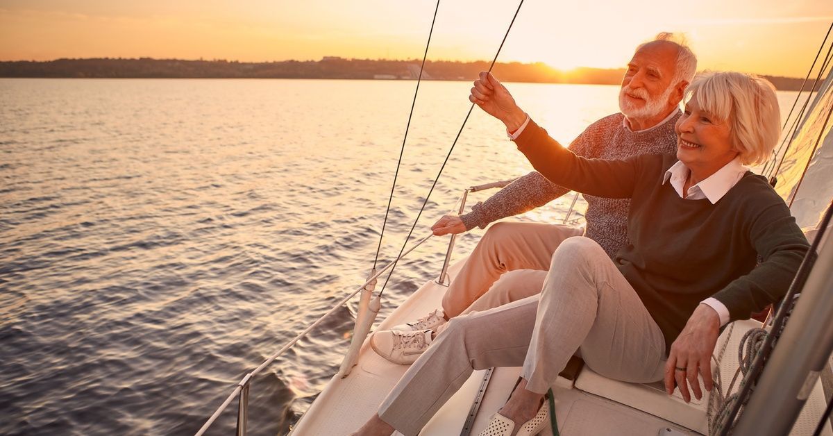 A senior couple relaxes on a boat as the sun goes down behind them. They're smiling and looking out at the water.