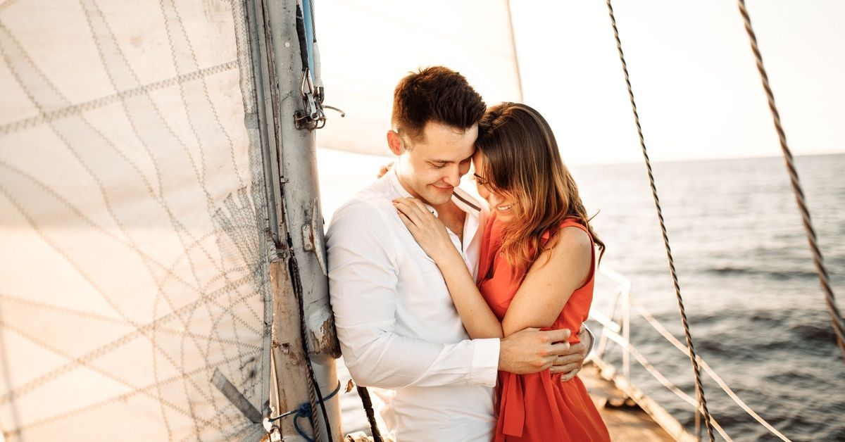 A man leans on a sailboat's mast and hugs a woman in an orange dress. They're smiling and looking down at their feet.