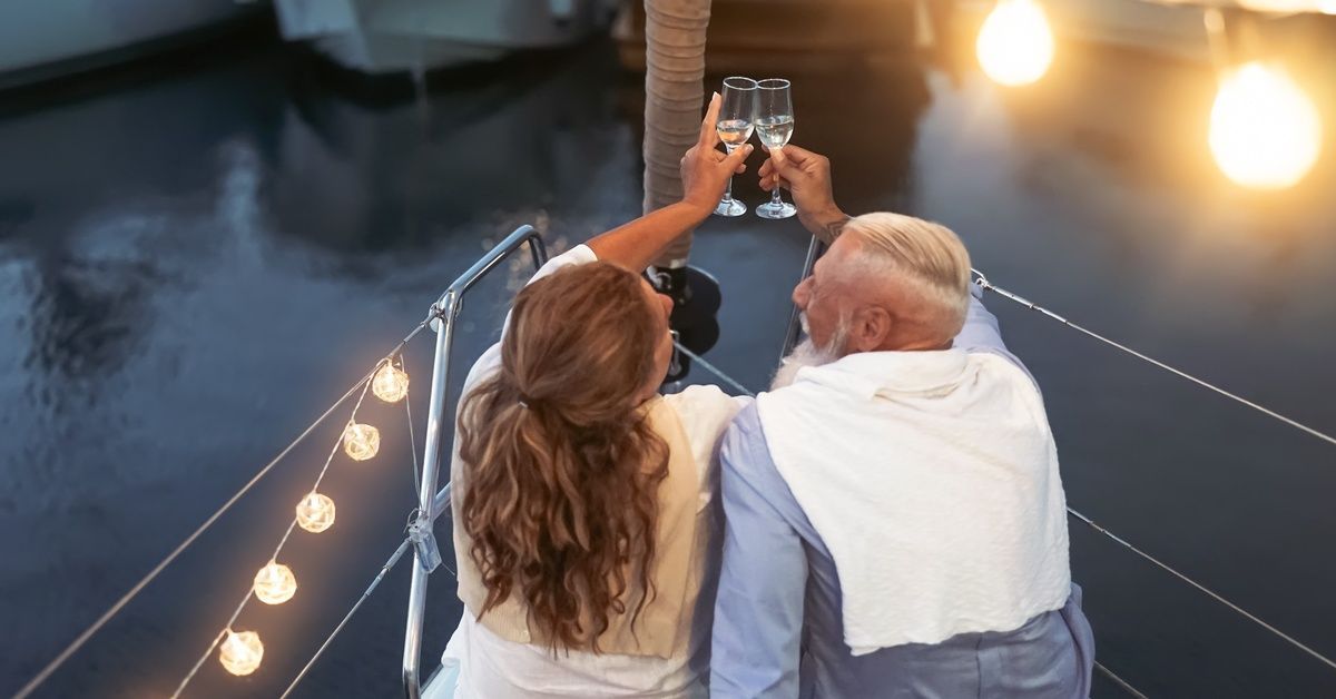 A man and woman sit on a boat's bow and clink champagne glasses together.