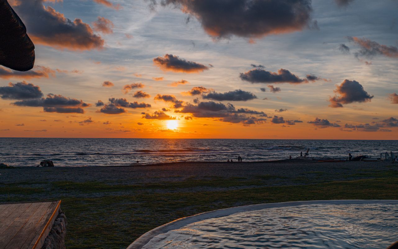 Sunset over the ocean with orange sky, scattered clouds, and a grassy shoreline in the foreground