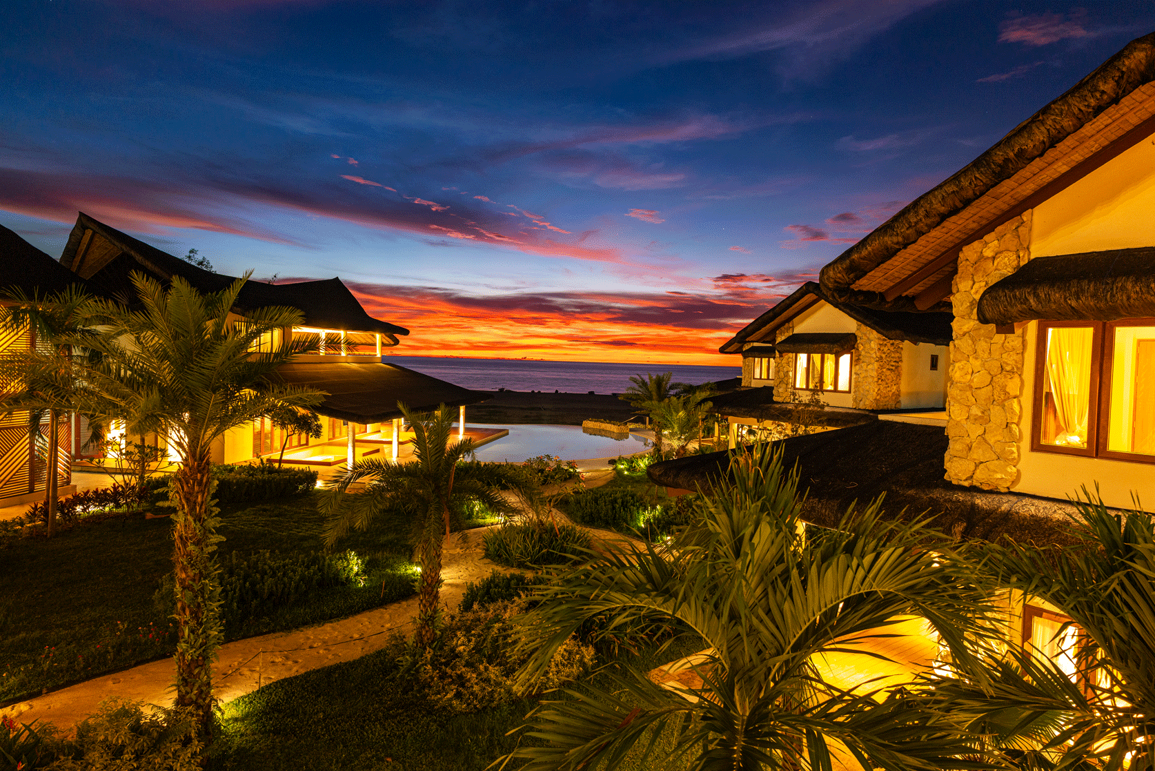 Tropical resort at sunset with lit villas, palm trees, and a glowing sky over the water