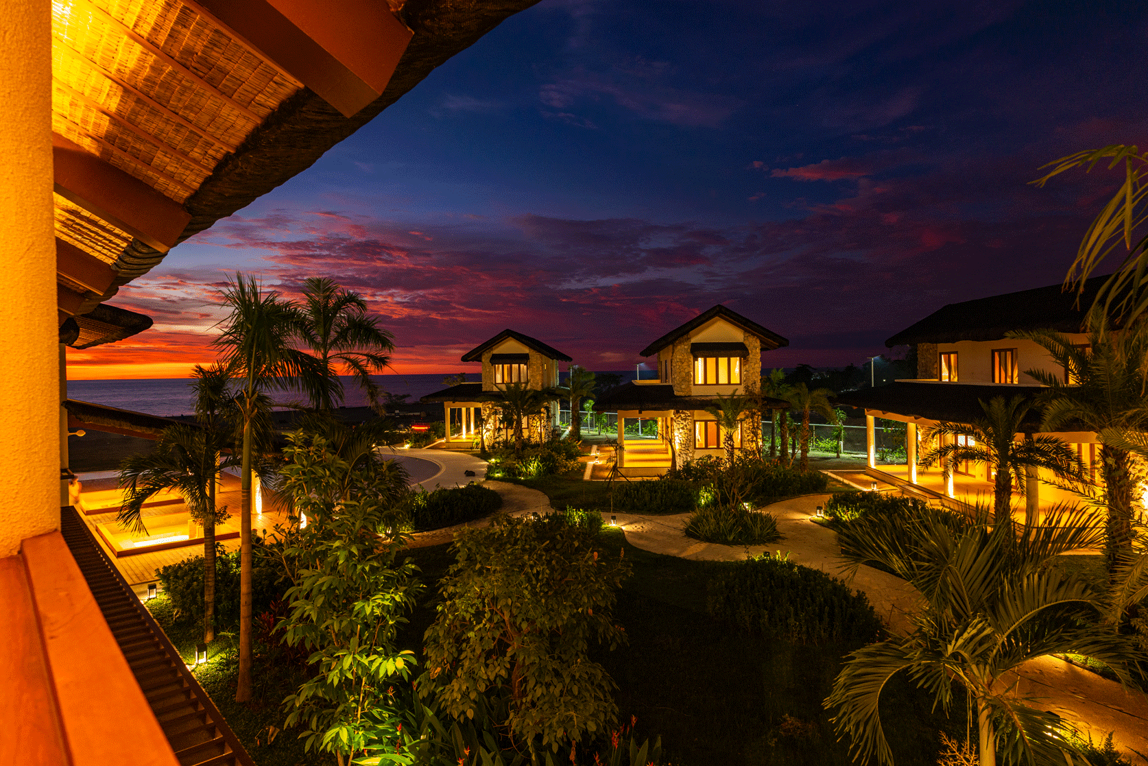 Tropical resort at sunset with lit villas, palm trees, and a vibrant purple-orange sky