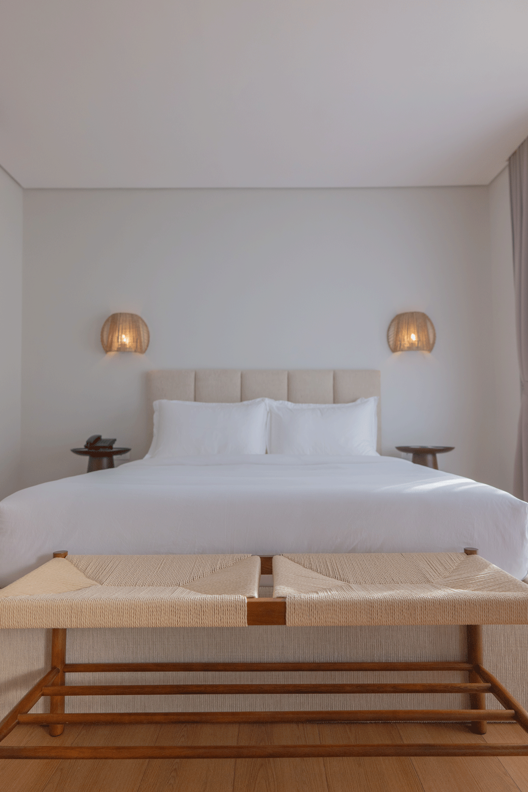 Minimalist hotel room with king bed, rattan sconces, and woven bench