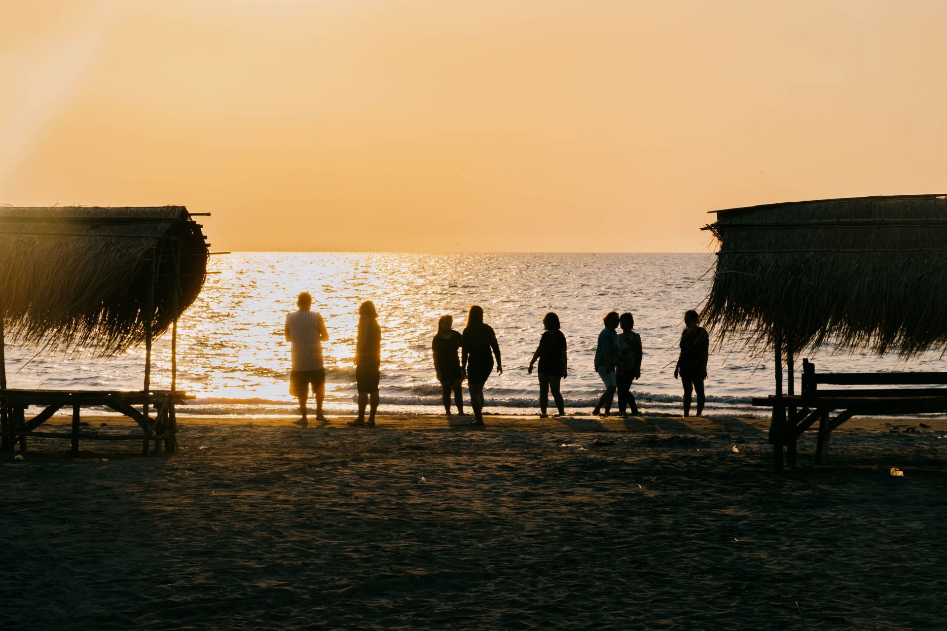 Silhouetted group standing on a beach at sunset, facing the ocean between two thatched huts