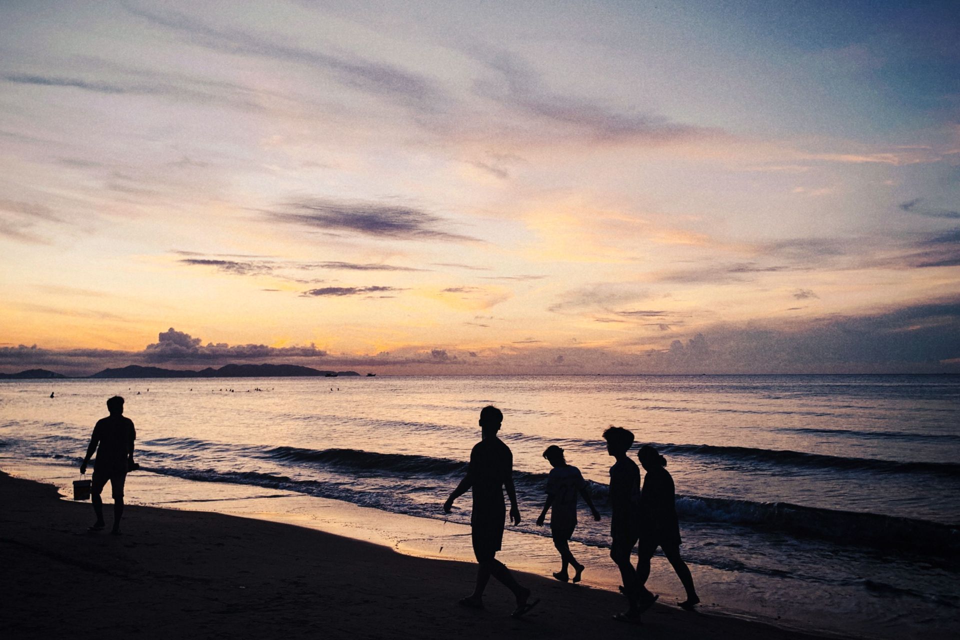 Silhouetted people walking along a beach at sunset, with waves and a colorful sky.