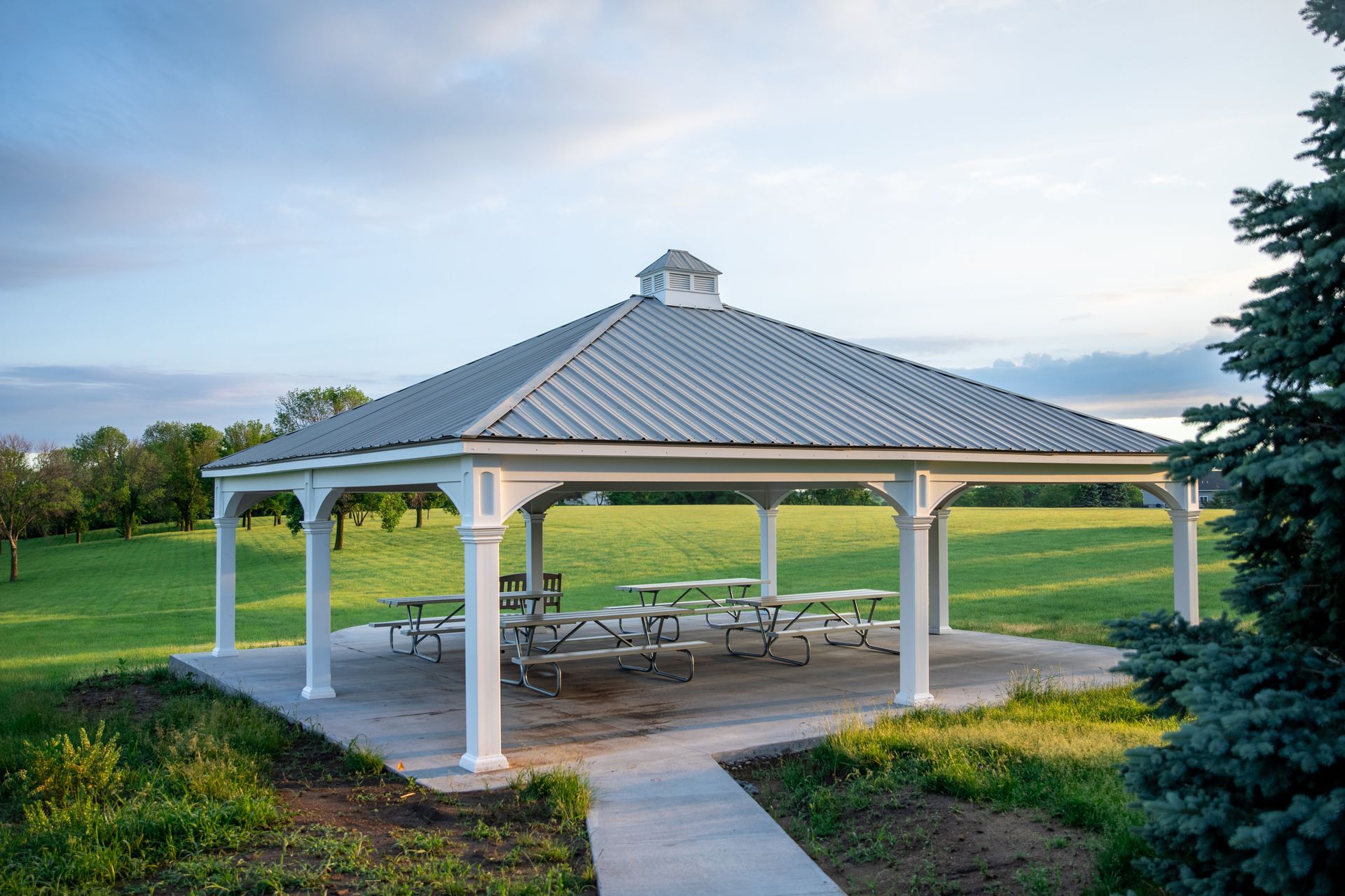 A white gazebo with a metal roof and picnic tables in a park.