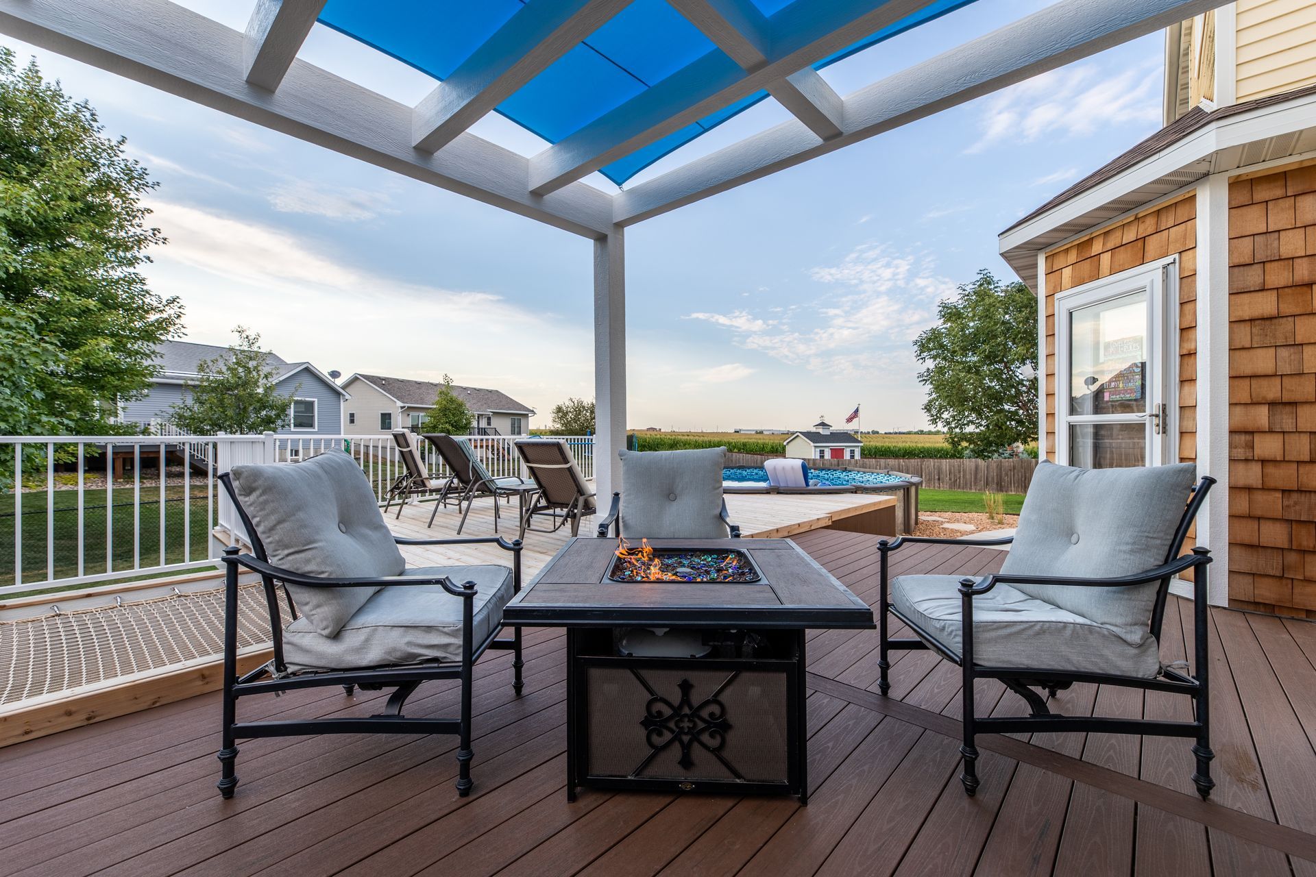 A wood deck with a fire pit and chairs under a blue pergola.