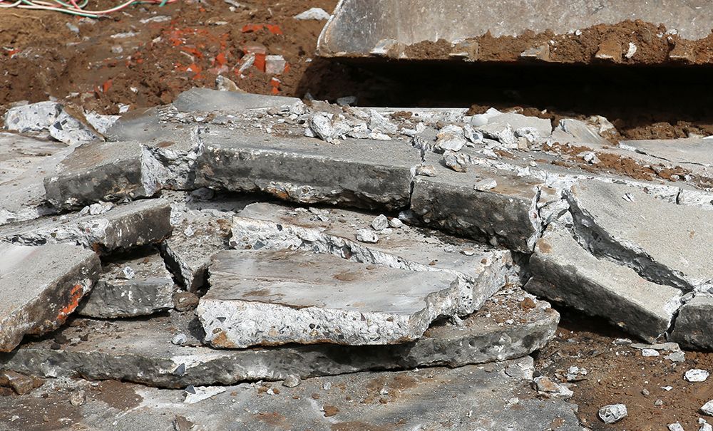 A pile of broken concrete slabs resting on brown dirt, with a portion of heavy machinery visible in the background.