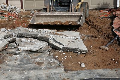Skid steer removing broken concrete slabs from a driveway. Dirt, red bricks, and a sledgehammer visible.