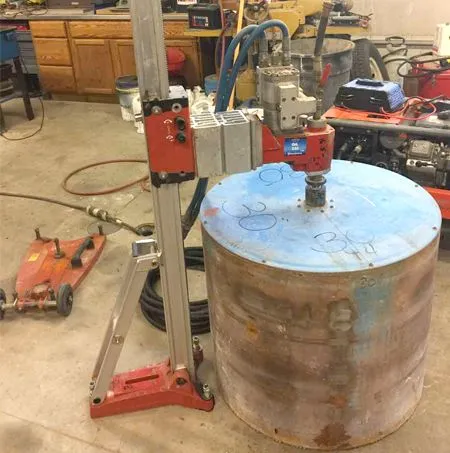 A core drilling machine cutting a circle from a large, cylindrical metal container in a workshop.